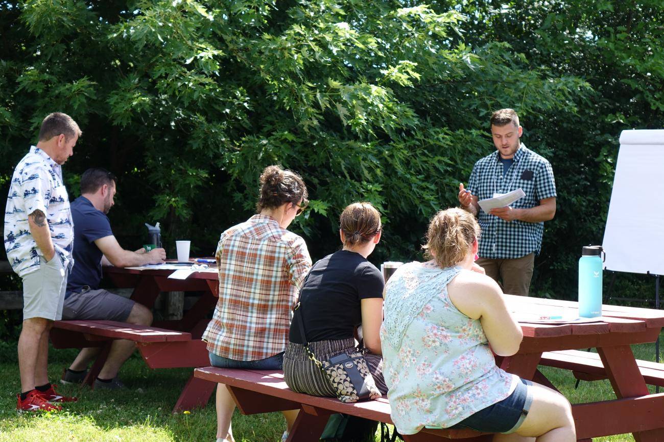 A man is standing in front of a small group of seated people in conversation outside at picnic tables at the Mabee Farm Historic Site. 